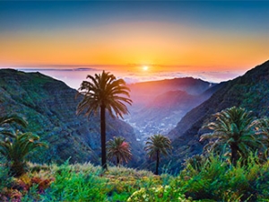 Beautiful view of amazing tropical scenery with exotic palm trees and mountain valleys above wide open sea in golden evening light at sunset with blue sky and clouds in summer, Canary Islands, Spain