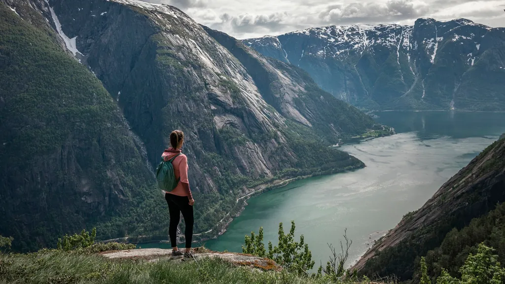 Ein Mädchen steht auf einem Felsvorsprung und blickt auf den majestätischen Fjord von Eidfjord in Norwegen. Umgeben von hohen, grünen Bergen und ruhigem Wasser, wirkt die Landschaft ruhig und beeindruckend. Der Himmel ist leicht bewölkt mit Sonne, die die Szene sanft beleuchtet.
