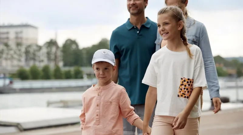 Eine fröhliche Familie spaziert entlang eines Flussufers. Im Vordergrund halten ein Junge mit heller Kappe und ein Mädchen mit geflochtenem Zopf lächelnd Händchen. Dahinter gehen ihre Eltern entspannt und zufrieden. Die Szene vermittelt Urlaubsstimmung und gemeinsame Familienzeit in einer idyllischen Umgebung.