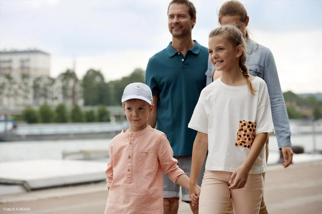 Eine fröhliche Familie spaziert entlang eines Flussufers. Im Vordergrund halten ein Junge mit heller Kappe und ein Mädchen mit geflochtenem Zopf lächelnd Händchen. Dahinter gehen ihre Eltern entspannt und zufrieden. Die Szene vermittelt Urlaubsstimmung und gemeinsame Familienzeit in einer idyllischen Umgebung.