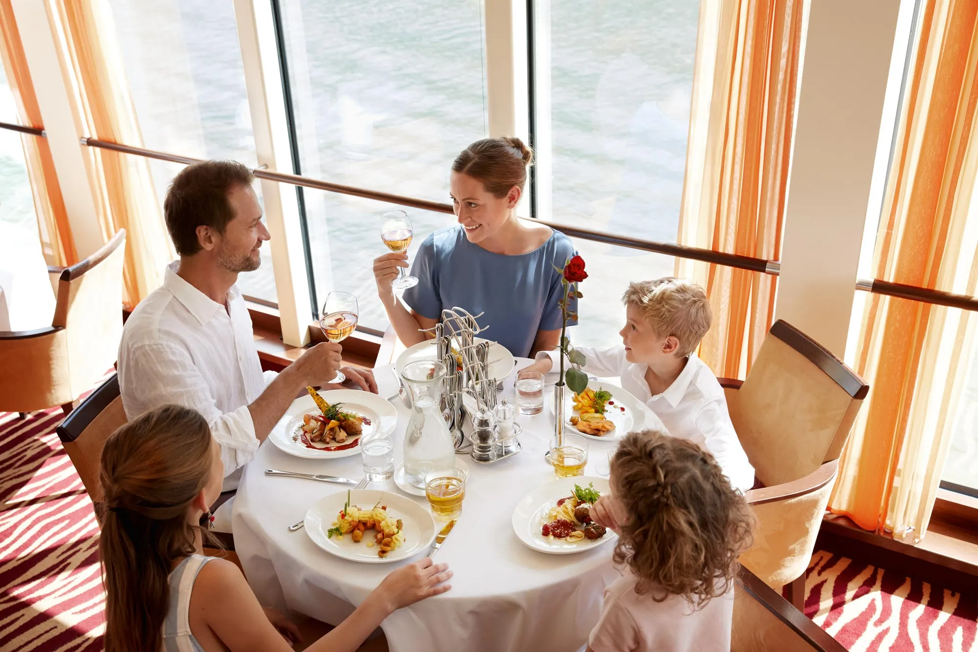 Eine Familie mit vier Personen sitzt an einem elegant gedeckten Tisch in einem Restaurant mit Panoramafenstern und Blick auf das Wasser. Die Eltern stoßen mit Weingläsern an und lächeln sich an, während die beiden Kinder ihr Essen genießen. Die Teller sind mit feinen Speisen angerichtet, und in der Mitte des Tisches steht eine dekorative Rose. Die warmen Farbtöne der Vorhänge und die stilvolle Einrichtung schaffen eine einladende Atmosphäre.