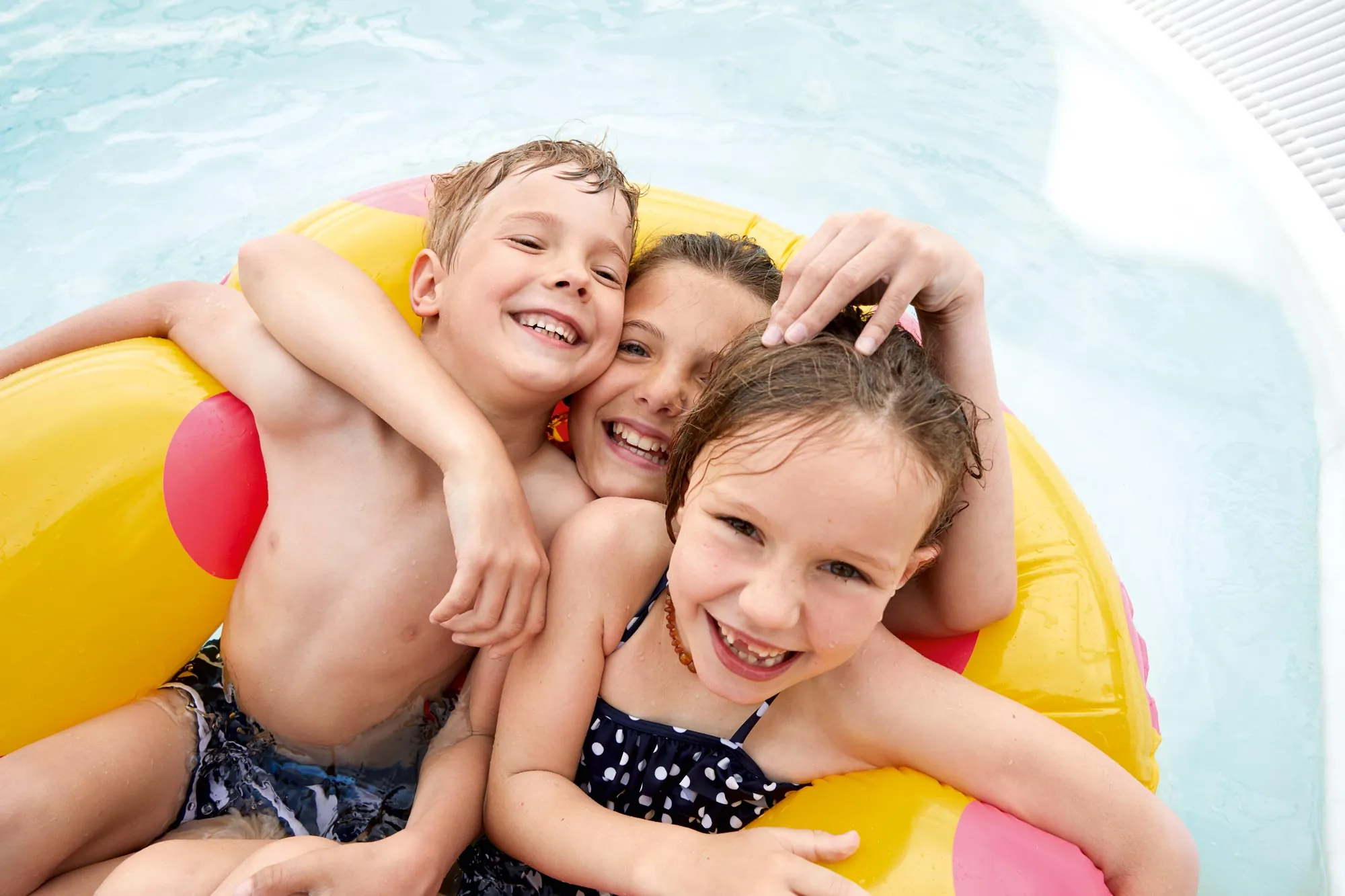 Drei lachende Kinder spielen gemeinsam in einem Pool und halten sich an einem großen, gelben Schwimmreifen fest. Ein Junge mit nassen Haaren umarmt zwei Mädchen – eines trägt einen dunkelblauen Badeanzug mit weißen Punkten, das andere ist teilweise hinter ihnen versteckt. Sie alle haben sichtlich Spaß im Wasser. Im Hintergrund ist das sprudelnde Wasser des Whirlpools zu sehen.