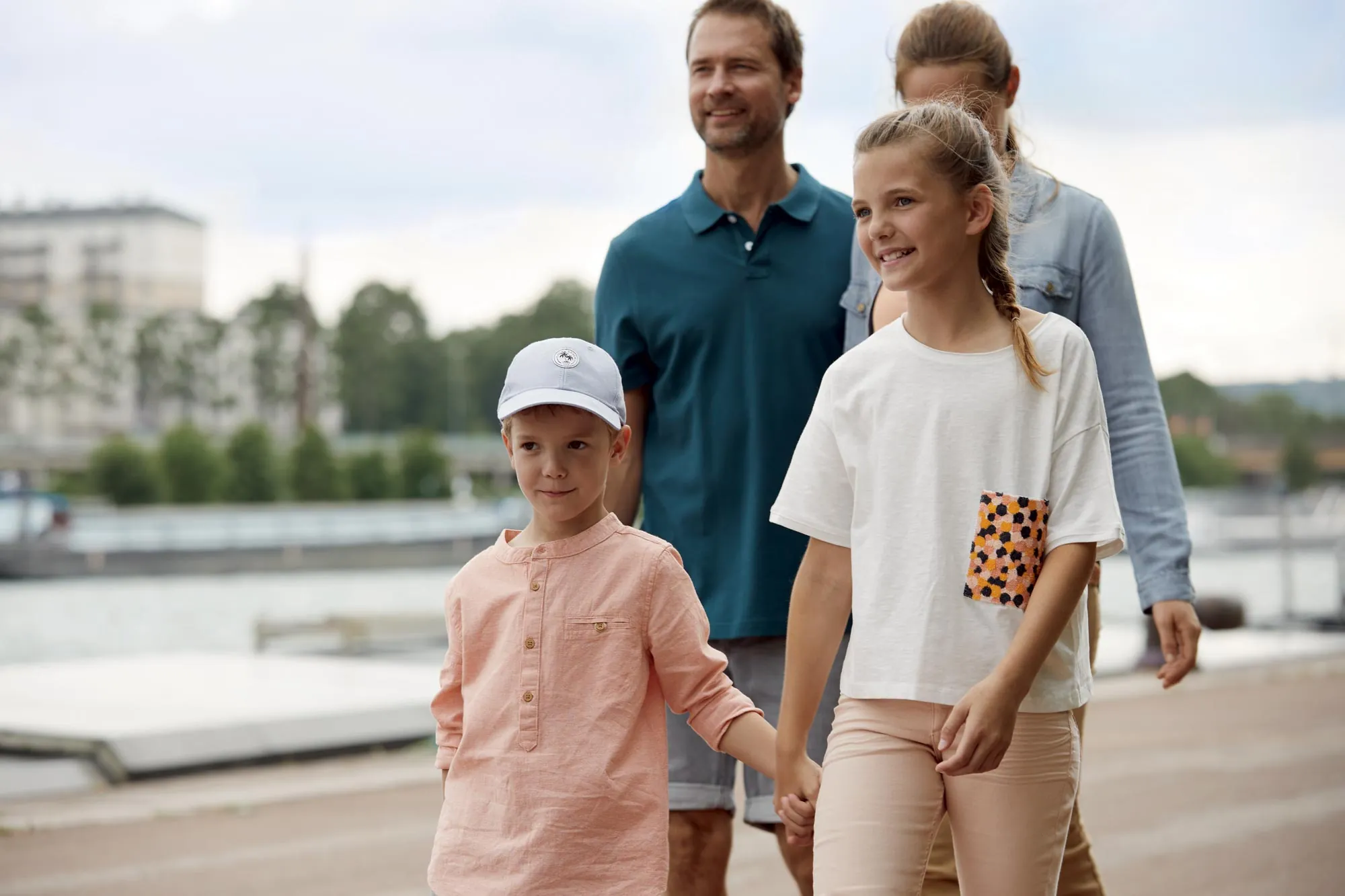 Eine fröhliche Familie spaziert entlang eines Flussufers. Im Vordergrund halten ein Junge mit heller Kappe und ein Mädchen mit geflochtenem Zopf lächelnd Händchen. Dahinter gehen ihre Eltern entspannt und zufrieden. Die Szene vermittelt Urlaubsstimmung und gemeinsame Familienzeit in einer idyllischen Umgebung.