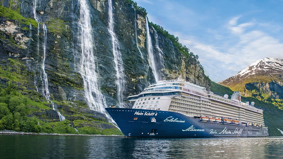 Das Kreuzfahrtschiff 'Mein Schiff 4' fährt durch eine beeindruckende Fjordlandschaft. Im Hintergrund ragen steile Felswände mit mehreren Wasserfällen empor, umgeben von grüner Vegetation. Schneebedeckte Berge und ein blauer Himmel vervollständigen die malerische Kulisse.