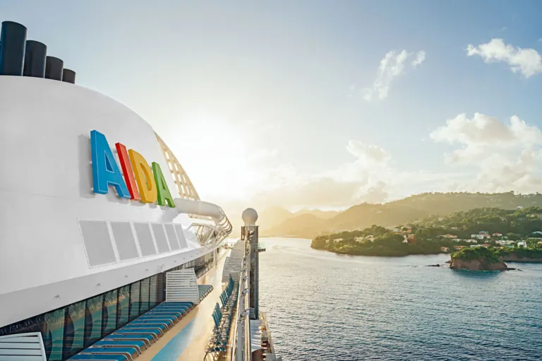 AIDA Kreuzfahrtschiff bei Sonnenaufgang vor tropischer Inselkulisse – Blick vom Oberdeck auf das Meer und eine grüne Küstenlandschaft.
