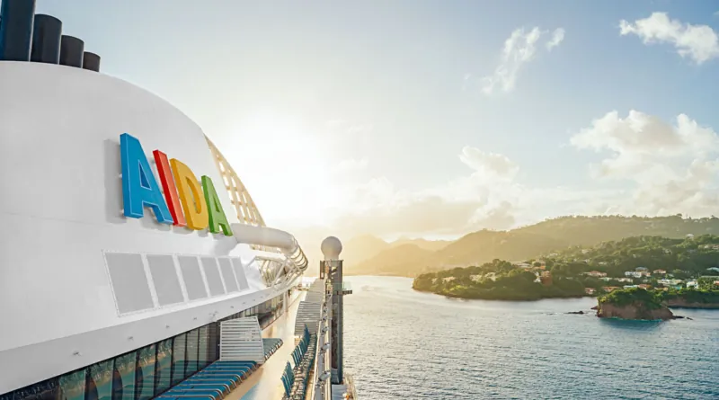AIDA Kreuzfahrtschiff bei Sonnenaufgang vor tropischer Inselkulisse – Blick vom Oberdeck auf das Meer und eine grüne Küstenlandschaft.