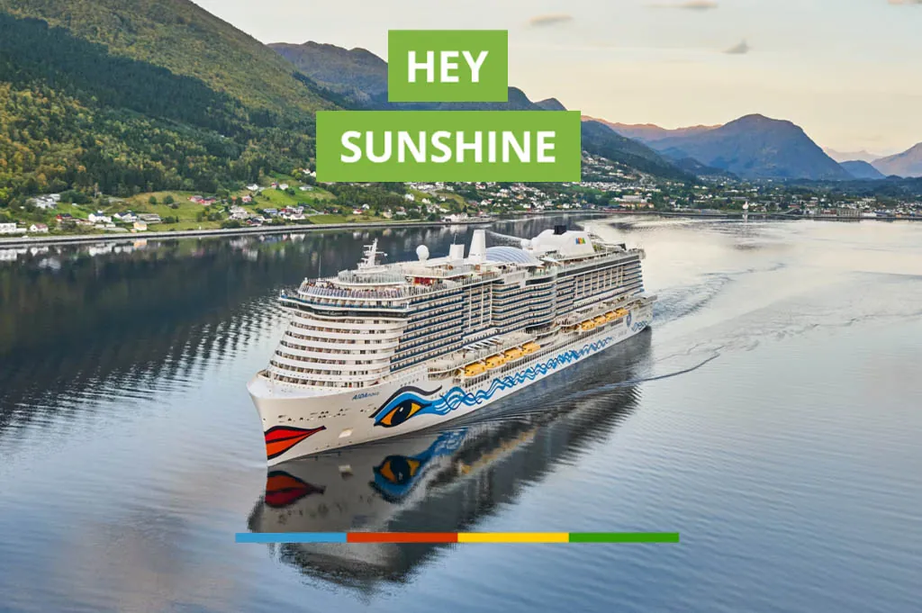 Ein großes Kreuzfahrtschiff der AIDA-Flotte fährt durch einen ruhigen Fjord, umgeben von grünen Bergen und einer kleinen Ortschaft am Ufer. Über dem Schiff steht in grünen Kästen der Schriftzug „HEY SUNSHINE“. Am unteren Bildrand befindet sich ein farbiger Balken in Blau, Rot, Gelb und Grün.