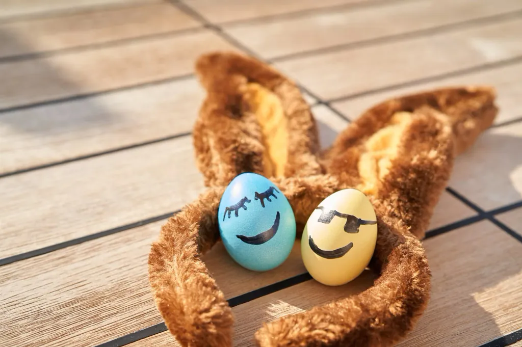 Zwei bunt bemalte Ostereier mit lachenden Gesichtern – eines blau mit geschlossenen Augen und Wimpern, das andere gelb mit Sonnenbrille – sitzen nebeneinander in einem braunen Plüschhasenohren-Stirnband auf einem Holzboden auf einem Kreuzfahrtschiff in der Sonne.