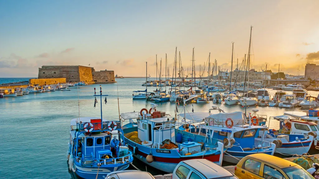 Stimmungsvoller Sonnenuntergang über dem Hafen von Heraklion auf Kreta mit bunten Fischerbooten, Segeljachten und der venezianischen Festung Koules im Hintergrund.
