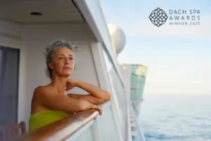 Eine Frau in einem grünen Handtuch lehnt entspannt am Balkon eines AIDA Kreuzfahrtschiffs, rechts oben das Logo „DACH Spa Awards Winner 2025“.