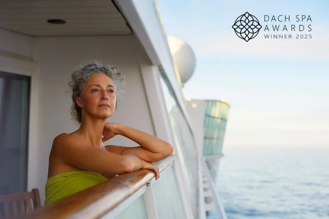 Eine Frau in einem grünen Handtuch lehnt entspannt am Balkon eines AIDA Kreuzfahrtschiffs, rechts oben das Logo „DACH Spa Awards Winner 2025“.
