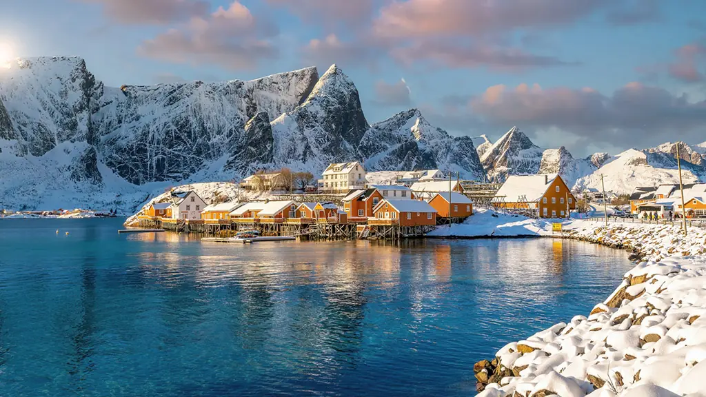 Winterliche Szenerie der Lofoten in Norwegen mit bunten Holzhäusern am Wasser, verschneiten Bergen und klarem blauen Himmel.