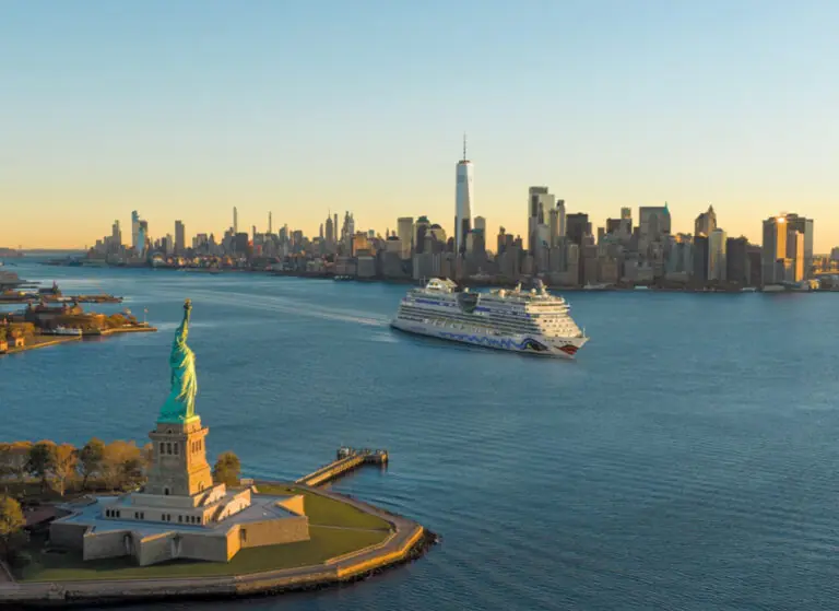 Luftaufnahme der Freiheitsstatue auf einer kleinen Insel im Hafen von New York, rechts daneben ein vorbeifahrendes Kreuzfahrtschiff auf blauem Wasser und dahinter die dicht bebaute Skyline Manhattans mit dem One World Trade Center bei tief stehender Sonne.
