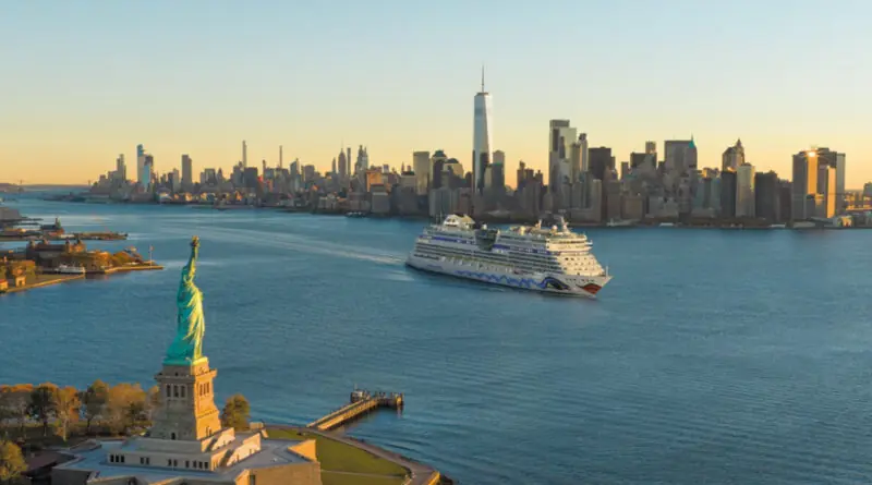 Luftaufnahme der Freiheitsstatue auf einer kleinen Insel im Hafen von New York, rechts daneben ein vorbeifahrendes Kreuzfahrtschiff auf blauem Wasser und dahinter die dicht bebaute Skyline Manhattans mit dem One World Trade Center bei tief stehender Sonne.