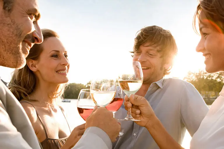 Vier Erwachsene stehen dicht beieinander im warmen Abendlicht und halten Weiß- und Roséweingläser zum Anstoßen hoch; im Hintergrund ist ein ruhiges Gewässer mit Bäumen zu sehen, die Szene wirkt sommerlich und freundlich.