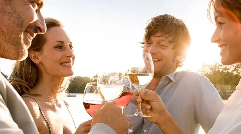 Vier Erwachsene stehen dicht beieinander im warmen Abendlicht und halten Weiß- und Roséweingläser zum Anstoßen hoch; im Hintergrund ist ein ruhiges Gewässer mit Bäumen zu sehen, die Szene wirkt sommerlich und freundlich.