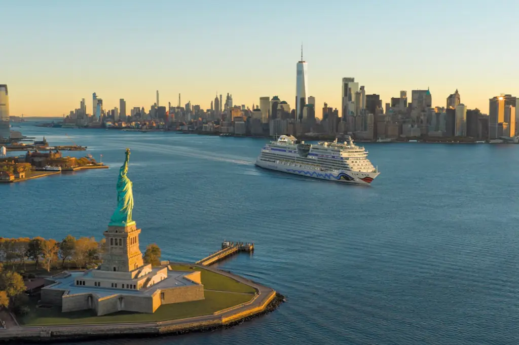 Luftaufnahme der Freiheitsstatue auf einer kleinen Insel im Hafen von New York, rechts daneben ein vorbeifahrendes Kreuzfahrtschiff auf blauem Wasser und dahinter die dicht bebaute Skyline Manhattans mit dem One World Trade Center bei tief stehender Sonne.