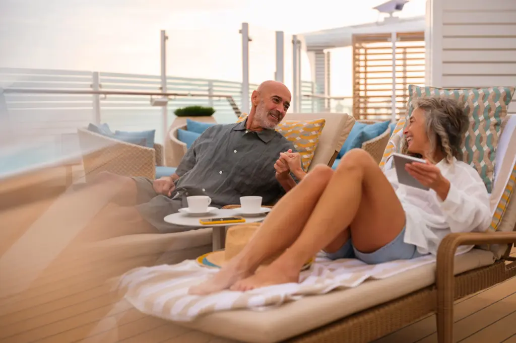 Älteres Paar liegt auf gepolsterten Sonnenliegen auf dem Außendeck eines Kreuzfahrtschiffes, mit Kissen, Kaffeetassen und Tablet, umgeben von modernem Deckmobiliar und hellem Tageslicht.