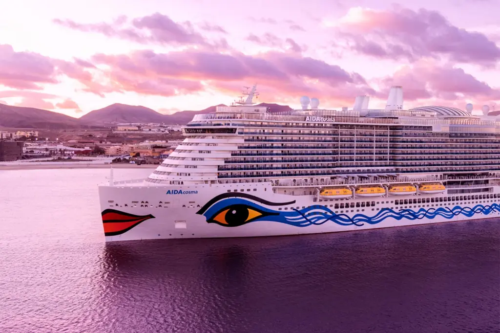 AIDAcosma Kreuzfahrtschiff mit typischem AIDA Kussmund fährt bei rosa-pinkem Sonnenuntergang vor einer Küste über das Meer, fotografiert im Querformat für romantische AIDA Kreuzfahrtangebote.