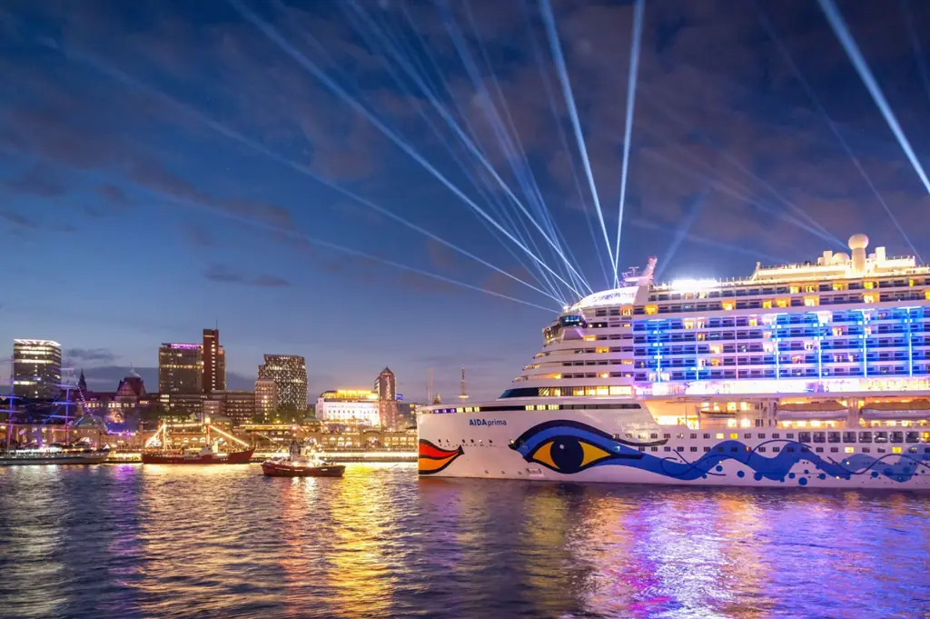 Großes Kreuzfahrtschiff AIDAprima mit typischem Kussmund-Design fährt bei Nacht beleuchtet durch den Hamburger Hafen, im Vordergrund spiegeln sich die Lichter auf der Elbe, im Hintergrund leuchtet die Skyline der Stadt.