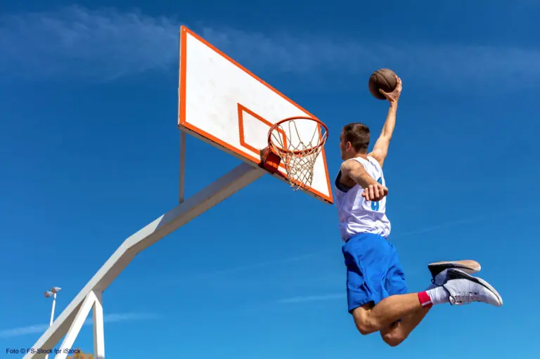 Junger Basketballspieler springt hoch vor blauem Himmel und dunkt den Ball kraftvoll in einen Outdoor-Korb.