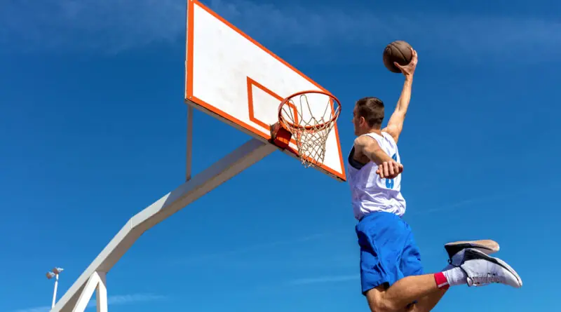 Junger Basketballspieler springt hoch vor blauem Himmel und dunkt den Ball kraftvoll in einen Outdoor-Korb.